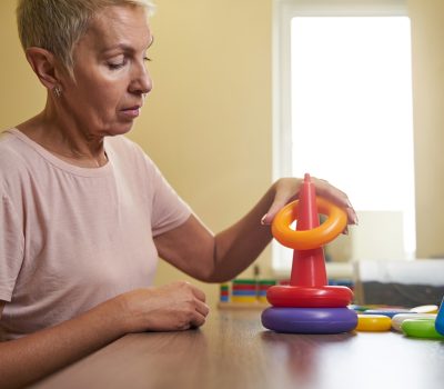 Caucasian woman performing recovery activity for motoric improvement by collecting colored toy pyramid in rehabilitation center